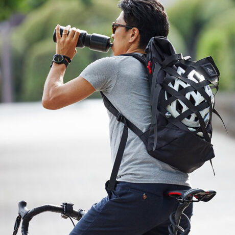 Man on a bicycle is drinking water and wearing the durable Braasi Wicker backpack, using the external net to hold his helmet.